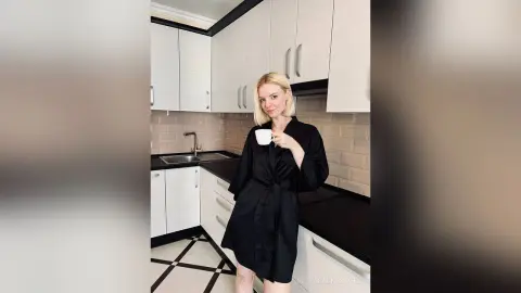 Blonde woman in a black silk robe enjoying coffee in a modern white and black kitchen.