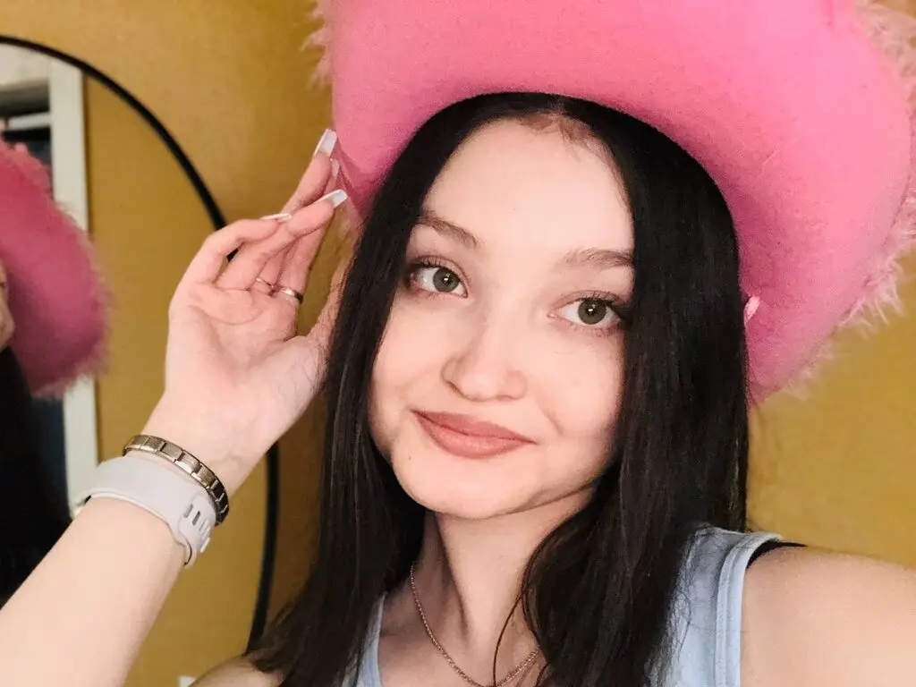 Young woman with long dark hair wearing a pink fuzzy hat and silver jewelry in a warm indoor setting.