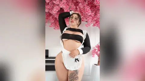 Curvy woman in a black and white maid outfit posing against pink cherry blossom flowers.