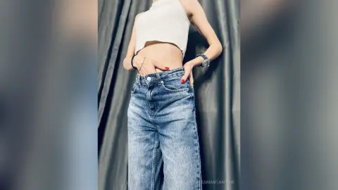 Young woman in a white crop top and high-waisted blue jeans posing confidently against a grey curtain backdrop.