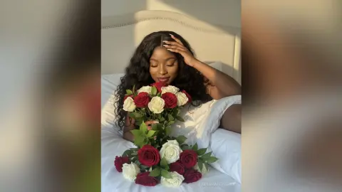 Smiling Black woman in white loungewear holding a large bouquet of red and white roses in a bright bedroom.