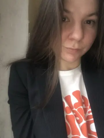 Young woman with long brown hair wearing a white graphic t-shirt and black blazer in a soft indoor setting.