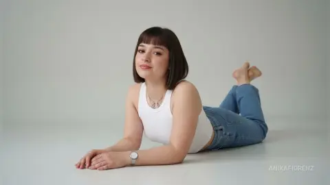 Smiling young woman in a white tank top and blue jeans lying on a white studio floor.