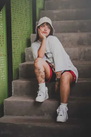 Trendy young woman in a white bucket hat and oversized t-shirt sitting on concrete stairs with a moody aesthetic.
