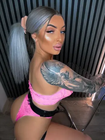 Woman with silver hair and large arm tattoo wearing pink lace lingerie in a modern setting.