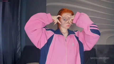MozelleRozycki wearing a pink and navy track jacket, holding her hands up near her face and framing her eyes with her fingers.