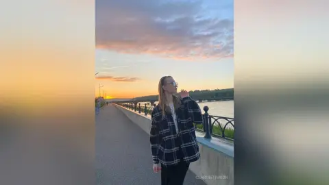 Young woman in a plaid flannel shirt standing on a pier during a golden sunset by the water.