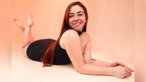 Smiling woman in a black bodysuit lying on a beige floor in a warm, minimalist studio setting.
