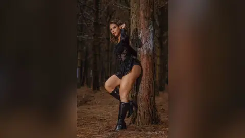 Glamorous woman in a black lace bodysuit and boots posing against a tree in a moody, dark forest setting.