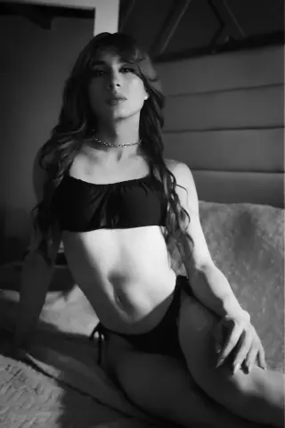 Young woman in black bikini lounging on a bed in a moody, black and white indoor setting.