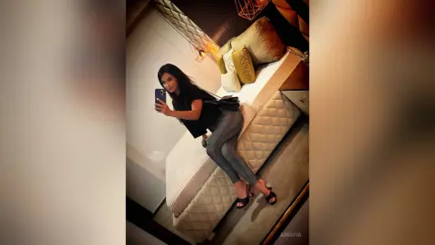 Stylish woman in black top and patterned trousers taking a selfie in a modern luxury bedroom.