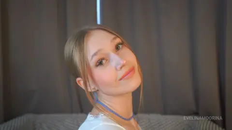 Smiling young woman in a white lace top and blue necklace posing against a dark curtain backdrop.