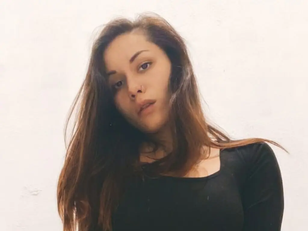 Young woman with long brown hair wearing a black top against a plain white background with a moody expression.
