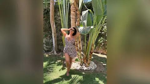 MissyJolie stands outdoors in a leopard print sundress, posing with her hands behind her head near tropical palm trees.