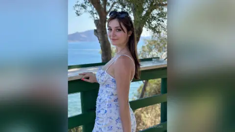Smiling woman in a white floral sundress relaxing on a balcony overlooking a scenic blue ocean view.