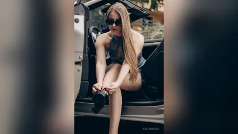 AdelinaKael wears sunglasses and a grey halter top while sitting in the open door of a vehicle, showing her long blonde hair and legs.