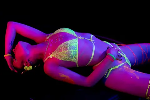 PaulaDiffiore lies on her back in a dark setting, wearing a yellow lace bra and matching undies with neon pink string details.