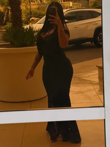 Woman in a sleek black midi dress taking a mirror selfie in a warm outdoor patio setting.