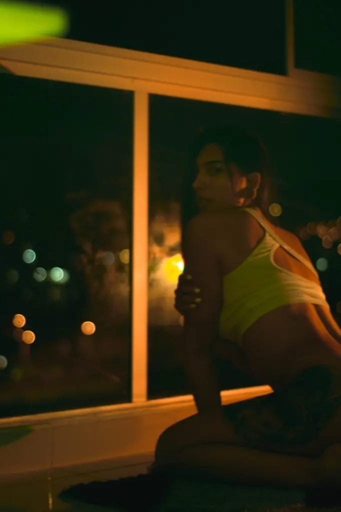 Louanebouroff — Young woman in a yellow crop top sitting by a window at night with warm ambient lighting.