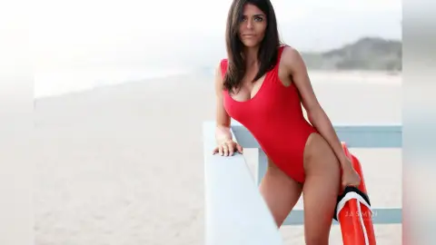 Beautiful woman in a red one-piece swimsuit posing on a sunny beach with a lifeguard buoy.