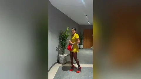 Stylish woman in a yellow mini dress and red heels posing in a modern minimalist hallway with indoor plants.