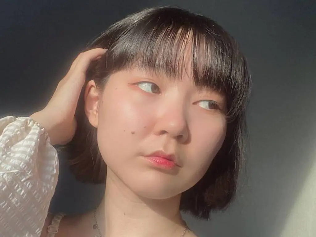 Young woman with short dark hair and bangs wearing a white textured blouse in soft natural sunlight.