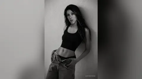 Moody black and white portrait of a young woman in a black tank top and denim jeans leaning against a wall.