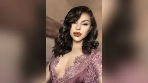 Glamorous woman with wavy dark hair wearing a mauve lace cocktail dress in a soft, elegant indoor setting.