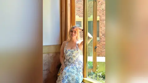 Smiling blonde woman in a blue floral sundress standing by a bright window in a sunny building.