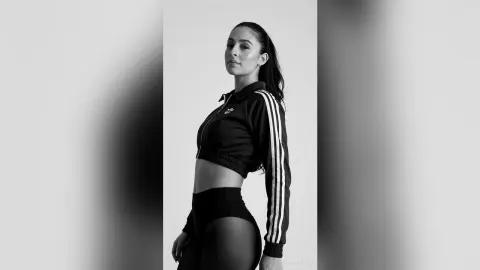 Confident woman in black Adidas tracksuit posing in a minimalist studio setting, black and white portrait.