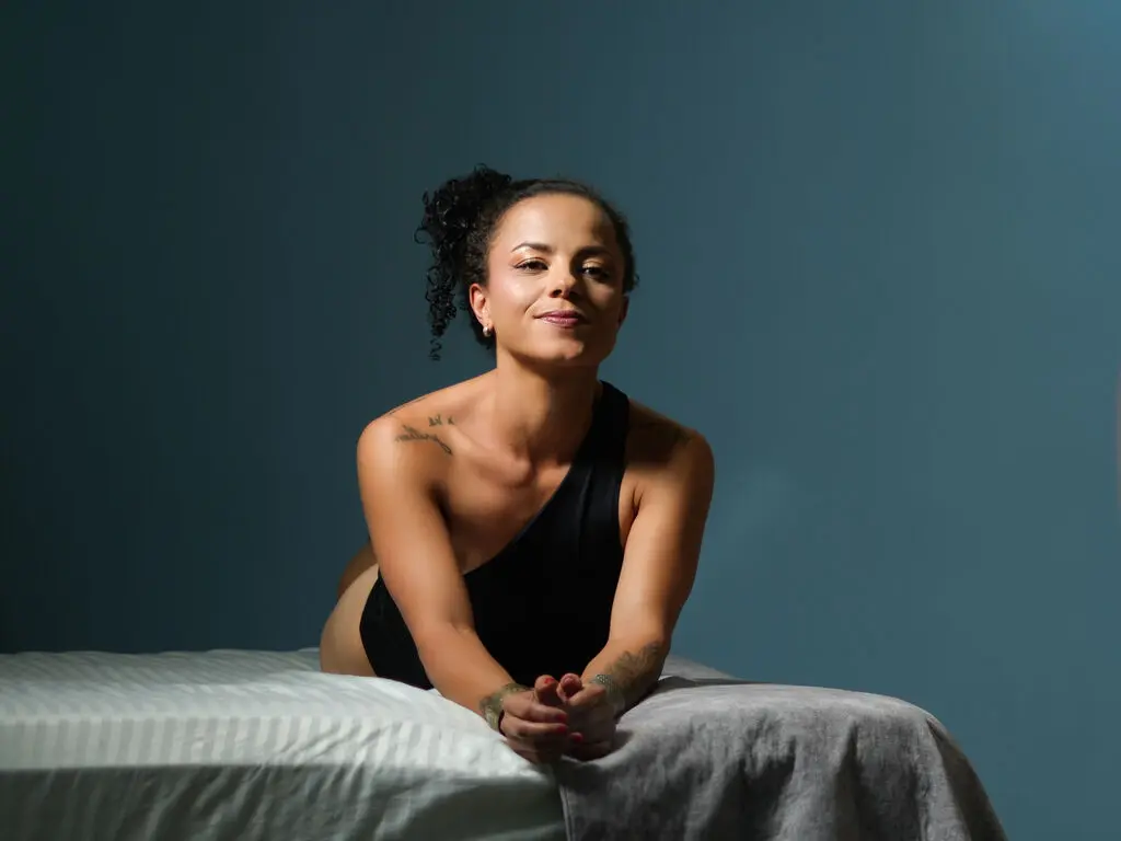 Smiling woman in a black tank top sitting on a bed against a dark blue background in soft studio lighting.