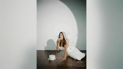 Elegant woman in a white dress sitting on a dark floor with a small white cake in a minimalist studio setting.