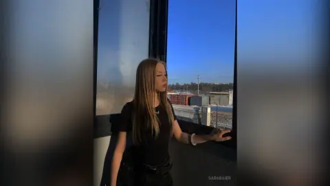 SaraBaier stands by a window in a black top, looking out at a view of buildings under a clear blue sky.