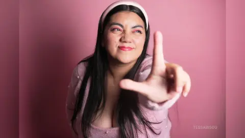 Cheerful woman in a pink sweater and headband posing playfully against a soft pink background.