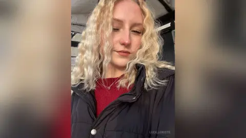 Blonde woman in a red sweater and black puffer jacket smiling softly in a car setting.