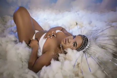 Glamorous woman in white lingerie and silver halo headpiece lying on soft white clouds in a dreamy setting.
