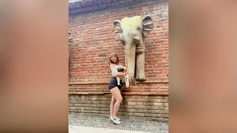 Young woman in a white crop top and black shorts standing by a large stone elephant statue against a brick wall.