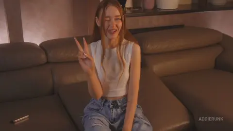 AddieRunk winks and sticks out her tongue while making a peace sign, wearing a white sleeveless crop top and light blue denim jeans while sitting on a brown leather couch.