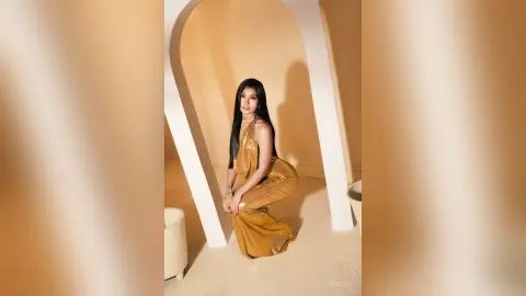 ElieMorgan poses in a long, shiny gold dress, sitting on her heels against a white arched background.