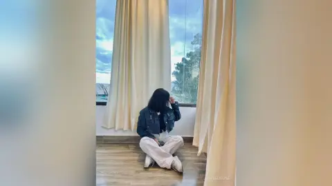 JanneMaine sits on a wood floor between cream-colored curtains, wearing a dark denim jacket and white pants.