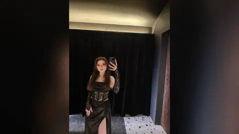 Young woman in a black lace bodysuit and satin skirt posing for a mirror selfie in a moody, dark room.