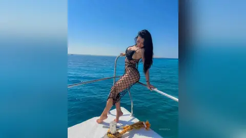 NicoleBella poses on a boat deck wearing a black fishnet monokini, showing off her long dark hair and bare skin against the blue ocean.