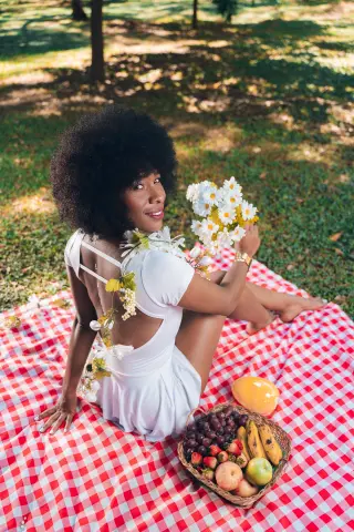 Smiling Black woman in a white summer dress holding daisies on a red checkered picnic blanket in a sunny park.