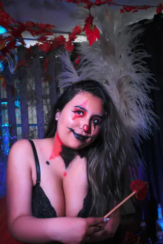 Smiling woman in spooky clown makeup and black lace outfit holding a red lollipop in a dark festive setting.