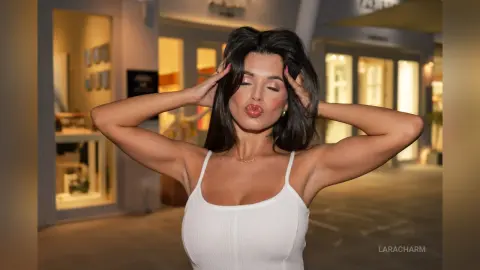 Young woman in a white ribbed tank top posing with hands in hair in a modern urban setting.