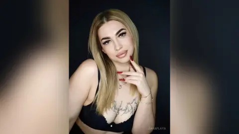 ZaraPlaya poses with her long blonde hair and red nails, wearing a black lace bra that shows off her chest tattoo against a dark background.