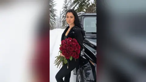 Elegant woman in black outfit holding large red rose bouquet in a snowy winter forest setting.