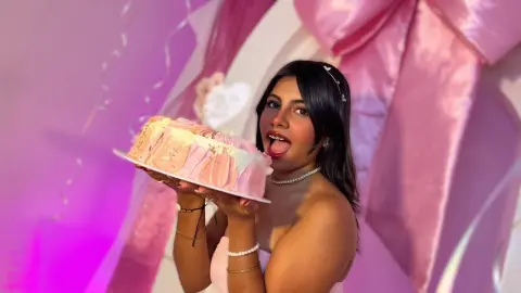 Cheerful woman in a white dress holding a pink frosted cake at a celebratory pink themed party.
