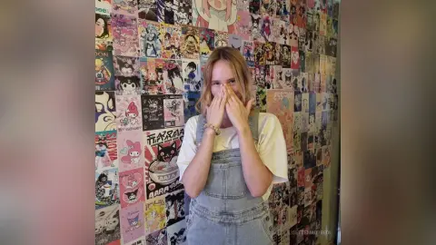 LisetteMaupredi wearing denim overalls and a white t-shirt, covering her mouth with both hands against a busy anime-style poster background.