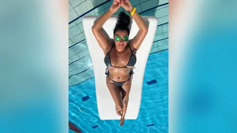 MissyJolie lies on a white lounge chair wearing a black string bikini and green sunglasses by a blue swimming pool.